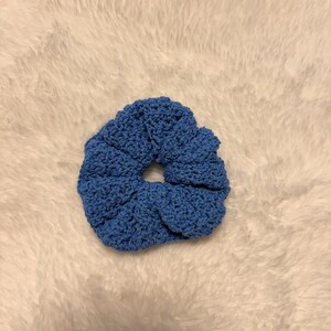 May include: A circular hair scrunchie made of blue yarn with a visible crochet stitch pattern. The accessory features a gathered, ruffled texture created by the elastic band inside, resulting in a soft, rounded shape with a central opening.