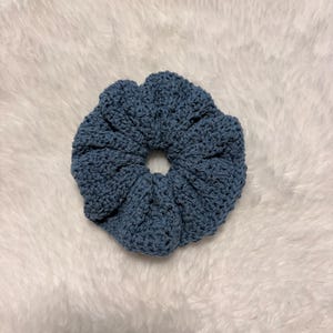 May include: A dusty blue hair scrunchie made of crocheted yarn. The accessory features a circular, gathered design with a visible textured stitch pattern throughout the fabric, creating a soft, ruffled appearance around the central elastic opening.