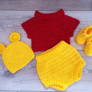 May include: A crocheted baby outfit in red and yellow. The outfit includes a hat with ears, a short-sleeved top, and bloomers.