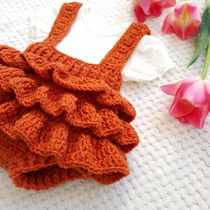 May include: A crocheted orange ruffled dress with straps. The dress is on a white fuzzy surface with pink tulips.