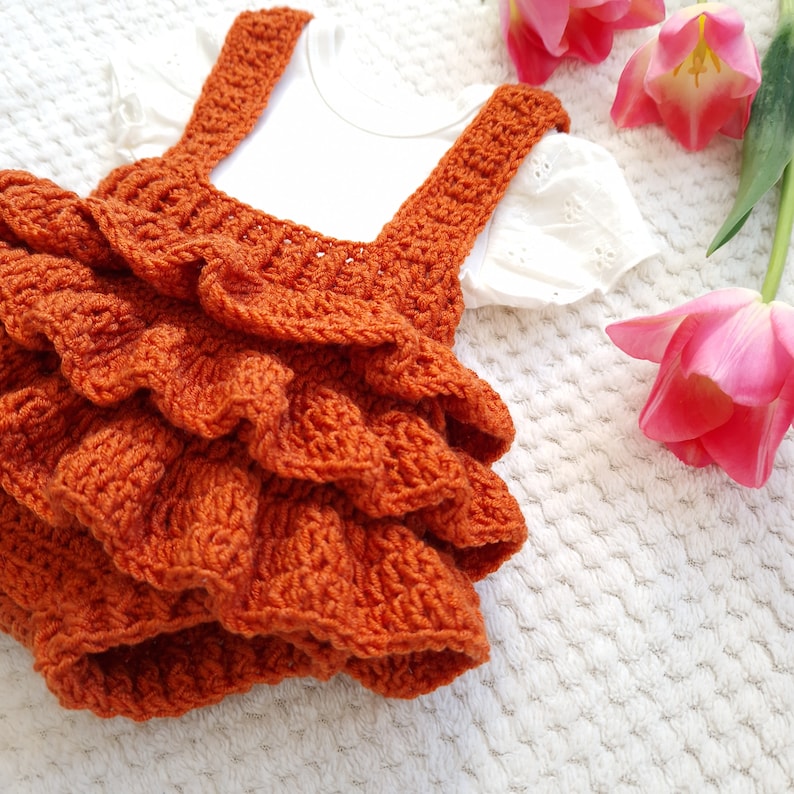 May include: A crocheted orange ruffled dress with straps. The dress is on a white fuzzy surface with pink tulips.