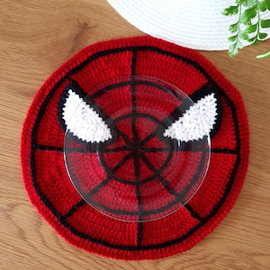 Puede incluir: Salvamanteles de ganchillo rojo y negro con un diseño de ojo de Spider-Man blanco. El salvamanteles es redondo y tiene un borde negro.