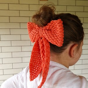 May include: A coral-colored crocheted bow hair tie. The bow is tied in a large, loose knot and has long, flowing tails.