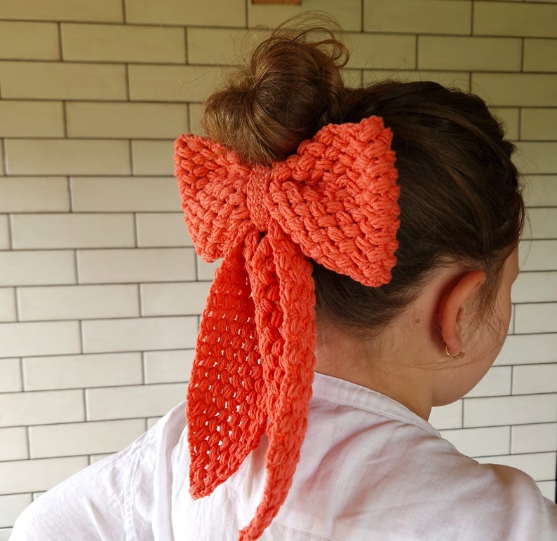 May include: A coral-colored crocheted bow hair tie. The bow is tied in a large, loose knot and has long, flowing tails.