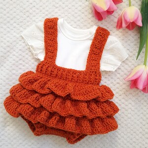 May include: A crocheted orange baby romper with a ruffled skirt and straps. The romper is on a white surface with pink flowers.