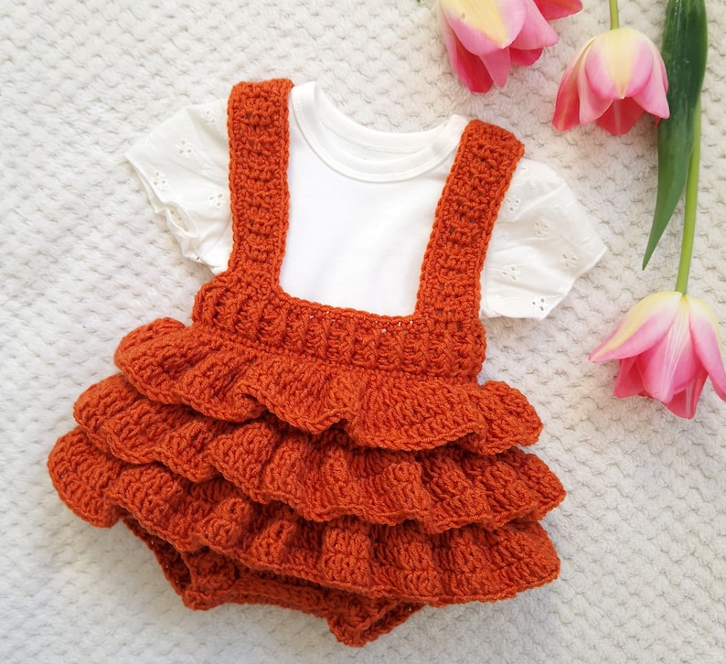 May include: A crocheted orange baby romper with a ruffled skirt and straps. The romper is on a white surface with pink flowers.