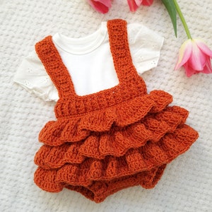 May include: A crocheted orange baby romper with a ruffled skirt and straps. The romper is made with a soft, textured yarn and has a simple, classic design.