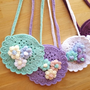 May include: Three crocheted purses with flower embellishments. The purses are in shades of green, purple, and white. Each purse has a crocheted strap.