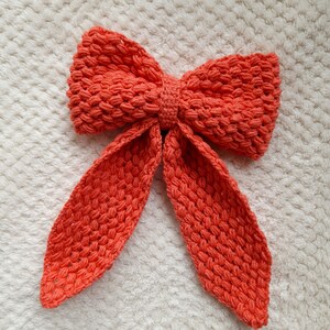 May include: A large, coral-colored crocheted bow with long tails. The bow is made with a textured stitch and is perfect for adding a touch of handmade charm to any project.