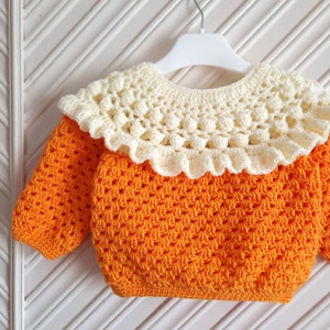 May include: A crocheted orange sweater with a white crocheted collar. The collar has a scalloped edge and a delicate pattern.