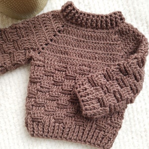 Può includere: Un maglione di lana all'uncinetto marrone con un motivo testurizzato. Il maglione ha un collo alto e maniche lunghe.