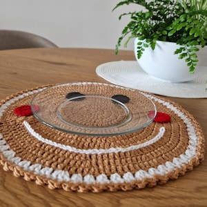 Może przedstawiać: Podkładka pod talerz wykonana na szydełku w kształcie piernikowego ludzika z białą obwódką i czerwonymi i czarnymi detalami. Na podkładce leży przezroczysty szklany talerz.