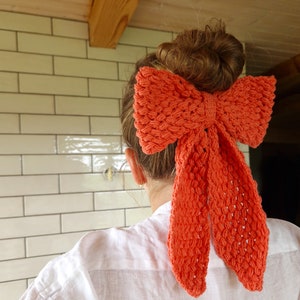 May include: A bright orange crocheted bow tied in a hair bow style. The bow is tied around a bun of brown hair.