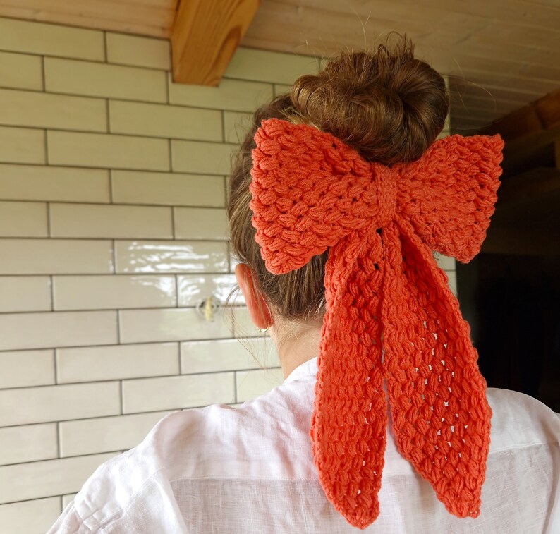 May include: A bright orange crocheted bow tied in a hair bow style. The bow is tied around a bun of brown hair.