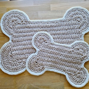 May include: Two handmade crochet dog bone-shaped mats. The mats are beige with a white border. The larger mat is approximately 61 cm long, and the smaller mat is about 30 cm long. The mats are made of a soft, textured yarn.