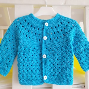 May include: A turquoise blue crocheted cardigan with white buttons. The cardigan has a simple design with a V-neck and long sleeves.