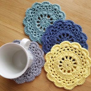Może przedstawiać: Trzy szydełkowe podkładki w kolorze niebieskim, szarym i żółtym. Podkładki są okrągłe i mają delikatny, koronkowy wzór.