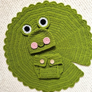 Puede incluir: Un conjunto de bebé de ganchillo verde hecho a mano, que incluye una manta, un gorro y una braguita. La manta tiene un borde festoneado y está diseñada para parecerse a una hoja de nenúfar. El gorro tiene mejillas rosas y la braguita tiene botones de madera.
