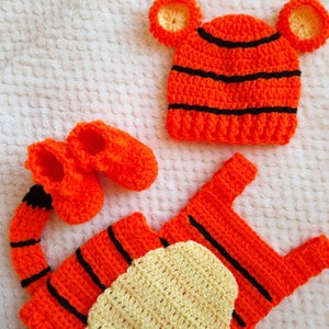 Könnte beinhalten: Ein gehäkeltes Tigerkostüm für ein Baby, bestehend aus einem orange-schwarz gestreiften Overall, einer passenden Mütze mit Ohren und Stiefeln.