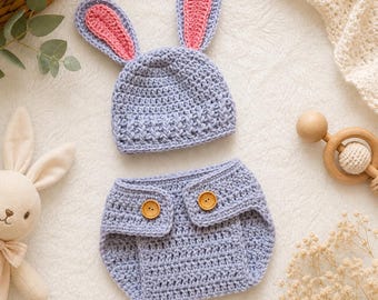 May include: A light blue crochet baby outfit, including a hat with pink-lined bunny ears and matching diaper cover with wooden buttons. The set is handmade and perfect for newborn photoshoots or baby showers.