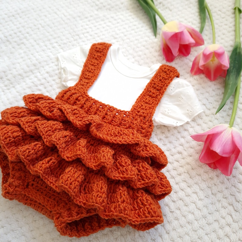 May include: A crocheted orange baby dress with a ruffled skirt and straps. The dress is laid out on a white textured surface with pink tulips.