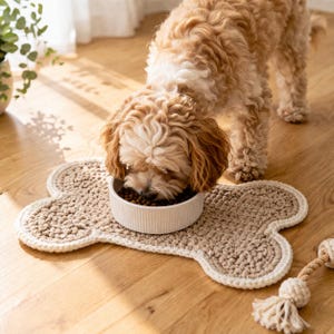 Könnte beinhalten: Ein kleiner, hellbrauner Hund frisst aus einem weißen Napf auf einer knochenförmigen, beigefarbenen Häkelmatte mit weißem Rand. Ein Seilspielzeug mit Quaste liegt in der Nähe. Der Hund hat lockiges, hellbraunes Fell.