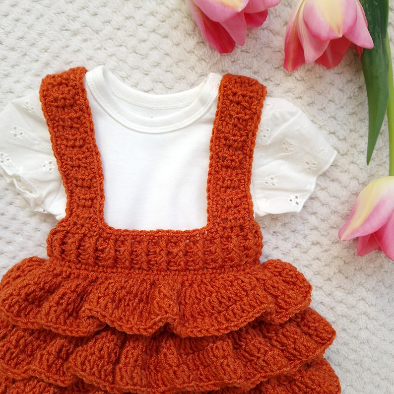 May include: A crocheted orange dress with a ruffled skirt and straps. The dress is layered with three ruffles. The dress is on a white fuzzy surface.