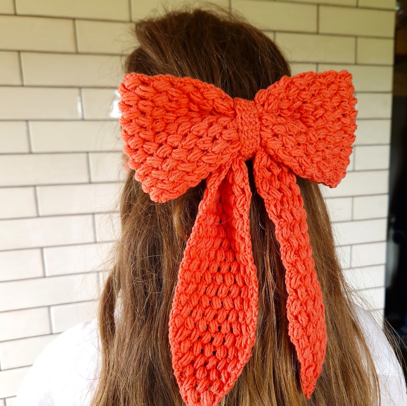 May include: A bright orange crocheted bow hair tie. The bow is tied in a large, loose knot with two long tails.