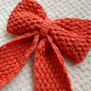 May include: A large, coral-colored crocheted bow. The bow is made of thick yarn and has a textured, bumpy surface.