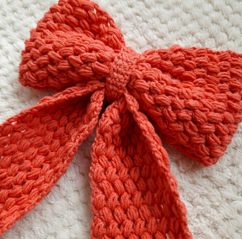 May include: A large, coral-colored crocheted bow. The bow is made of thick yarn and has a textured, bumpy surface.