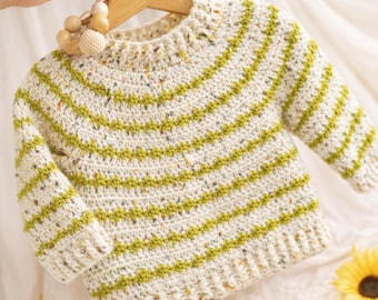 Op de afbeelding: Een crèmekleurige baby trui met lange mouwen en horizontale groene strepen. De trui is handgemaakt met een gespikkeld garen en heeft een ronde halslijn. Het wordt gepresenteerd op een houten hanger.