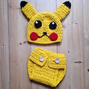 May include: A handmade yellow crochet Pikachu costume set, including a hat with black-tipped ears, black eyes, red cheeks, and a matching diaper cover with two white buttons. The set is designed for babies.