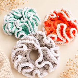 May include: Three handmade crochet scrunchies in shades of green, orange, and gray, each with a white trim. The scrunchies are arranged on a light beige surface, showcasing their textured, ruffled design. These hair accessories are perfect for adding a touch of handmade charm.