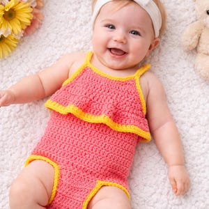 May include: A baby wearing a handmade pink crochet romper with yellow trim and a matching headband with a bow. The baby is smiling and lying on a white, textured surface. A teddy bear and flowers are in the background.