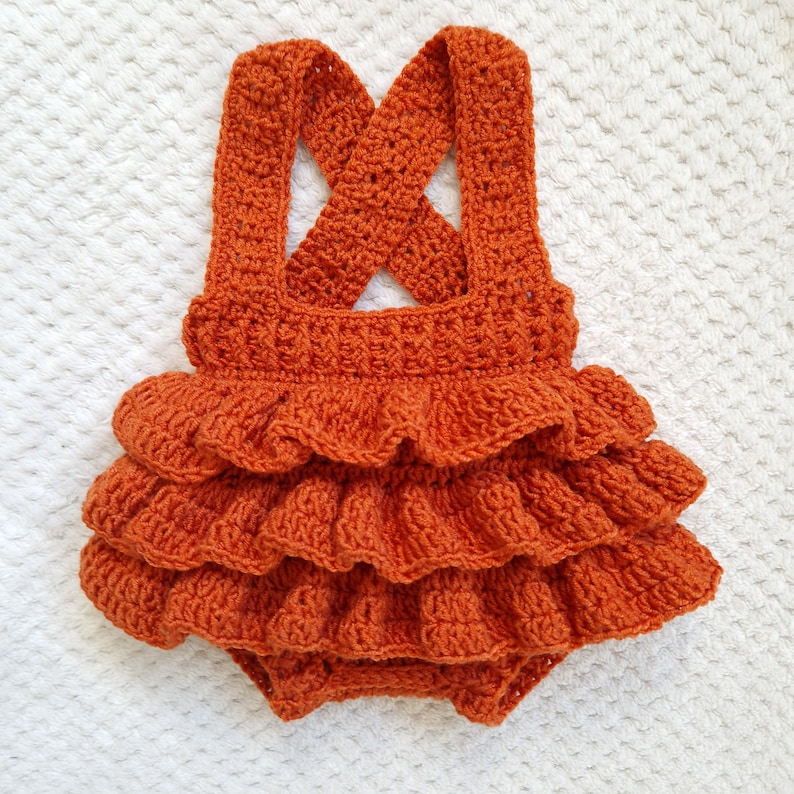 May include: A crocheted baby romper with a ruffled skirt in a burnt orange color. The romper has a criss-cross strap design in the back.