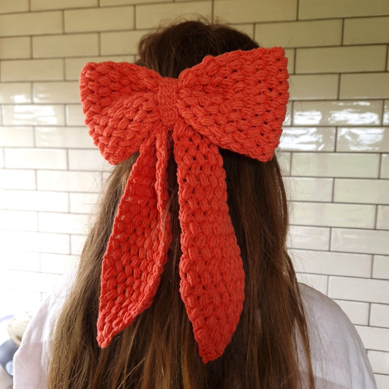 May include: A bright orange crocheted bow tied in the hair of a person with long brown hair.