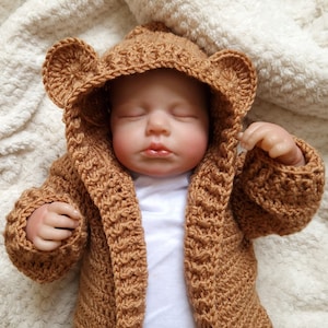 May include: A sleeping baby doll wearing a brown crocheted cardigan with a bear hood. The cardigan has a textured stitch pattern and the hood has bear ears.