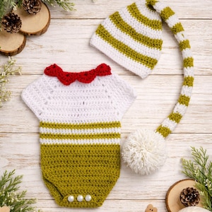 Op de afbeelding: Een handgemaakt gehaakt baby-outfit in wit en olijfgroen, met een romper met een rode geschulpte kraag en een bijpassende gestreepte hoed met een lang, bungelend uiteinde en een witte pompon. De outfit is versierd met drie parelmoer knopen.