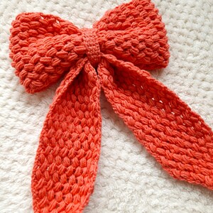 May include: A large, coral-colored crocheted bow with long tails. The bow is made of a textured yarn and has a soft, fluffy appearance.