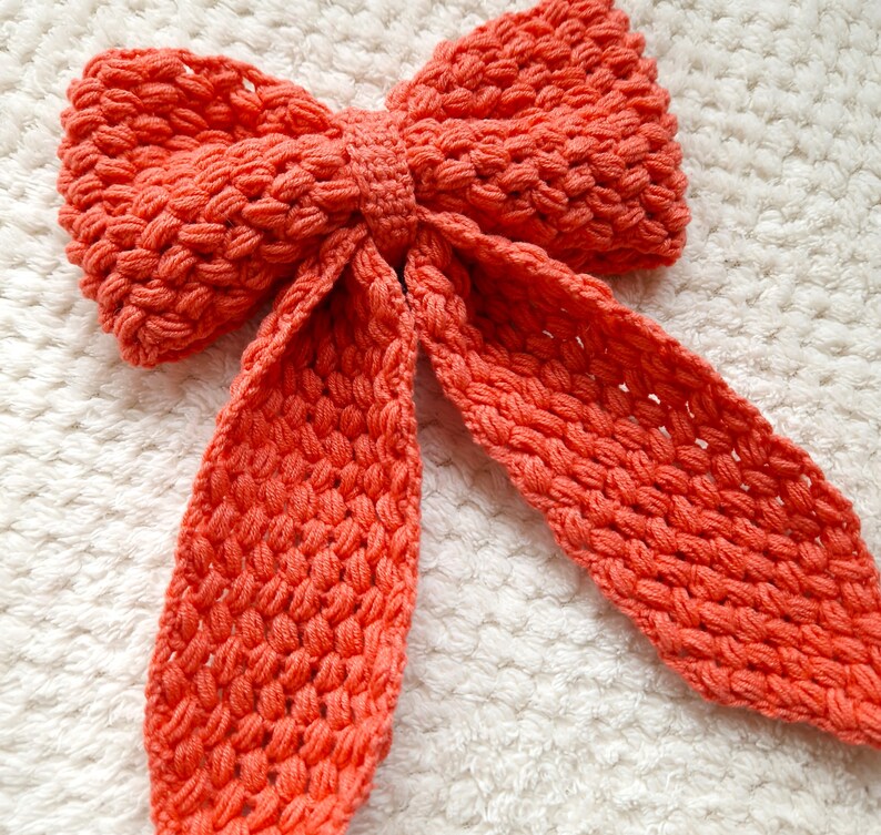 May include: A large, coral-colored crocheted bow with long tails. The bow is made of a textured yarn and has a soft, fluffy appearance.