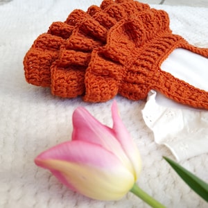 May include: A crocheted baby dress with a ruffled skirt in a burnt orange color. The dress is laid on a white fuzzy surface with a pink tulip.