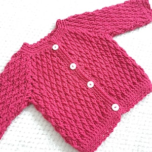 May include: A pink crocheted cardigan with white buttons. The cardigan has a ribbed texture and a classic button-up design.