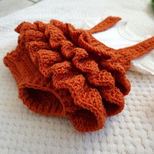 May include: A crocheted baby romper with a ruffled skirt in a burnt orange color.