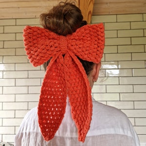 May include: A coral-colored crocheted bow hair accessory. The bow is tied in a large knot and has long tails that hang down the back.