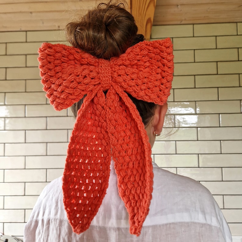 May include: A coral-colored crocheted bow hair accessory. The bow is tied in a large knot and has long tails that hang down the back.