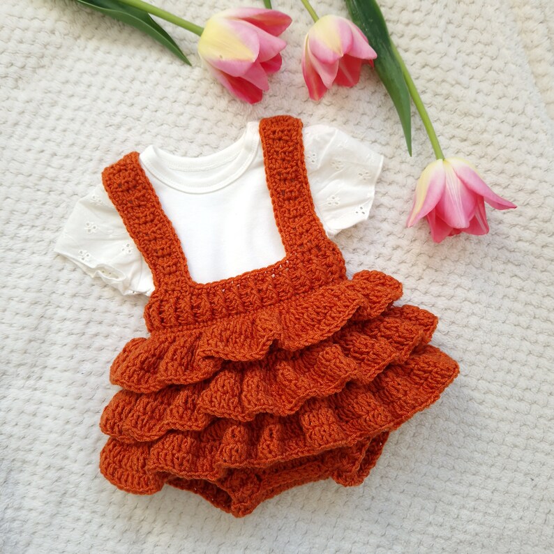 May include: A crocheted orange baby romper with a ruffled skirt and white sleeveless top.
