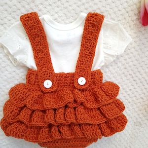 May include: A crocheted orange baby romper with white buttons and a ruffled skirt. The romper has straps that go over the shoulders.