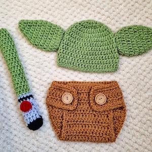 Peut inclure: Ensemble de vêtements pour bébé crocheté en vert, marron et bleu. L'ensemble comprend un chapeau avec des rabats en forme d'oreilles, une culotte avec des boutons en bois et un sabre laser. Le sabre laser est vert, bleu, rouge et noir.