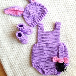 May include: Purple crocheted baby outfit with a matching hat and booties. The outfit features a tail and ears, resembling a donkey.