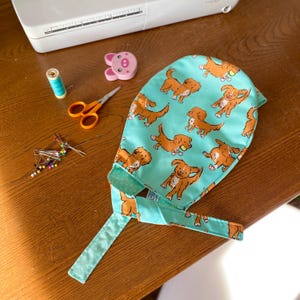May include: A light turquoise scrub cap featuring a repeating pattern of brown dogs in various playful poses. The cap has long fabric ties for securing it in place and is constructed from cotton fabric with a soft, smooth texture and finished edges.